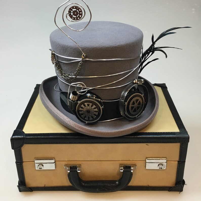 May include: A gray steampunk top hat with silver wire accents, a chain, and goggles. The hat features a black band and is adorned with a feather. It rests on a tan and black suitcase with a handle and silver clasps.