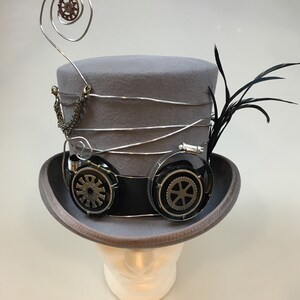 May include: A gray steampunk top hat with silver wire accents, a chain, and black goggles with gear details. A black feather and a decorative wire spiral with a gear are attached to the hat. The hat is displayed on a white mannequin head.