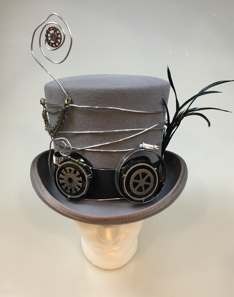 May include: A gray steampunk top hat with silver wire accents, a chain, and black goggles with gear details. A black feather and a decorative wire spiral with a gear are attached to the hat. The hat is displayed on a white mannequin head.