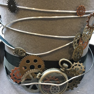 May include: A gray steampunk top hat with silver wire accents and decorative gears. The hat features a black ribbon band, a small pocket watch, and a metal tag with the number 45.