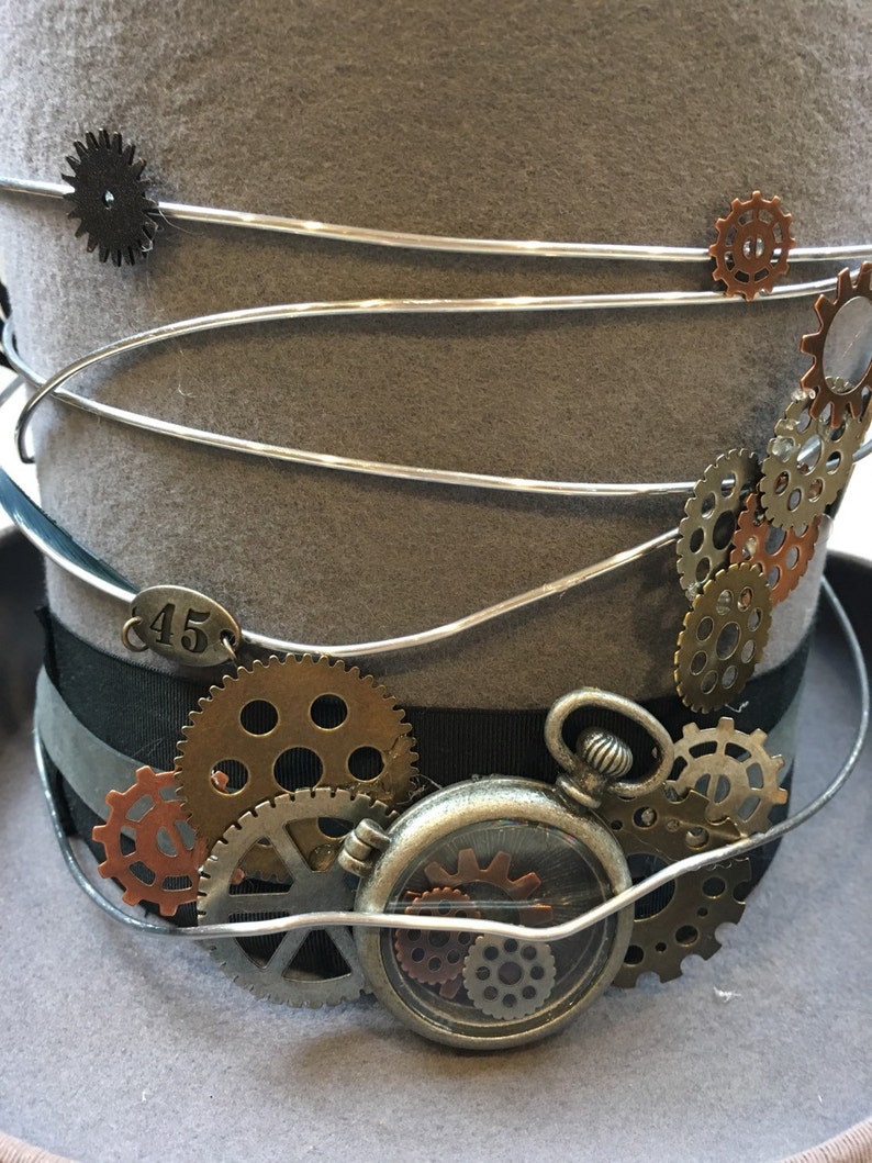 May include: A gray steampunk top hat with silver wire accents and decorative gears. The hat features a black ribbon band, a small pocket watch, and a metal tag with the number 45.