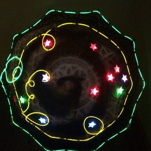 May include: A dark, umbrella-shaped object with a glowing green outline and yellow and green accents. The umbrella features several illuminated star-shaped lights in red, white, and green, creating a celestial design.