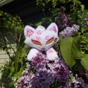 May include: White plush toy fox with pink and gold embroidered details. The fox is sitting on a bed of purple flowers.