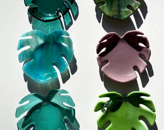 May include: Six glass monstera leaf shaped dishes in various colors, including green, blue, and pink. The dishes are all the same size and shape, and they are arranged in two rows of three.