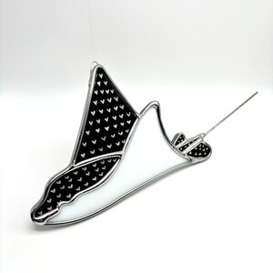 May include: A stained glass ornament shaped like a manta ray with a black and white pattern of hearts. The ornament is hanging from a silver string.