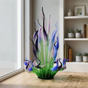 Vibrant Chihuly-style hand-blown glass sculpture featuring tall, flame-shaped glass reeds in green, cobalt blue, purple, and black hues, displayed in a bright modern living room corner next to a white bookshelf with succulents
