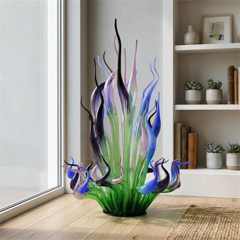 Vibrant Chihuly-style hand-blown glass sculpture featuring tall, flame-shaped glass reeds in green, cobalt blue, purple, and black hues, displayed in a bright modern living room corner next to a white bookshelf with succulents