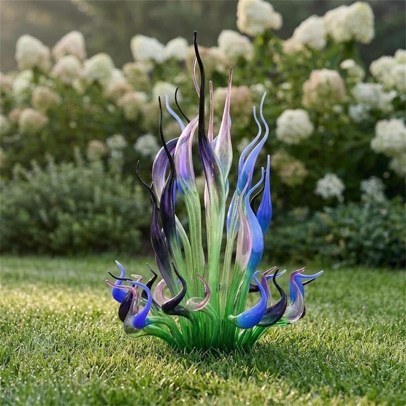Chihuly-style hand-blown glass sculpture with green, blue, and purple flame forms, displayed outdoors in a lush garden for patio or yard