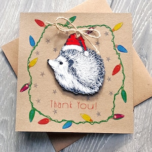 Tarjeta de agradecimiento navideña con regalo de erizo y adorno extraíble para el árbol. Adorno navideño de madera con forma de animal sobre una tarjeta de felicitación reciclada ilustrada.