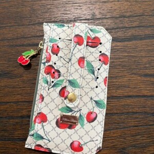 May include: A beige wallet featuring a repeating brown geometric pattern and a red cherry print with green leaves. The wallet includes a vertical strap with a gold-tone snap button, a side zipper with a cherry charm, and a small rectangular metal logo plate.