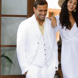 May include: A couple in white clothing standing in a doorway. The man is wearing a white linen suit and the woman is wearing a white linen dress with a wide-brimmed hat.