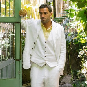 May include: A man wearing a white linen suit with a vest and a light yellow shirt. He is standing in front of a green door and a wooden walkway.