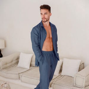 Pode incluir: Um conjunto de pijama de linho azul. O conjunto inclui uma camisa abotoada com bolso no peito e calças com cordão. O modelo está usando a camisa aberta. O pijama é de cor sólida e parece leve.