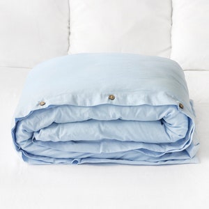 May include: A light blue linen-cotton blend duvet cover with three wooden buttons. The duvet cover is folded and placed on a white surface.