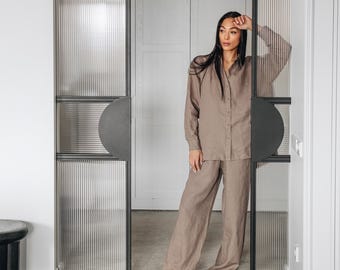 May include: A woman wearing a light brown linen shirt and matching pants stands in a doorway. The doorway features glass panels with vertical ribbed detailing and dark gray circular door handles. The floor has a dark wood herringbone pattern.