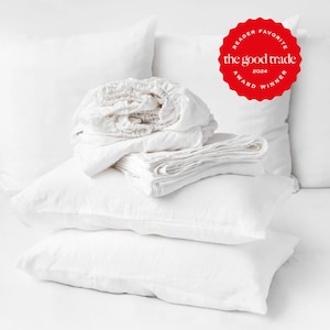 May include: A stack of white linen bedding including two rectangular pillows and folded sheets. A red circular badge in the upper right corner features the text "READER FAVORITE" and "the good trade" with "2024 AWARD WINNER" printed around the edge.