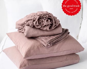 May include: A set of dusty rose linen bedding including a fitted sheet, a flat sheet, and two pillowcases stacked on a white bed. A red circular badge in the upper right corner features white text reading Reader Favorite The Good Trade Award Winner 2024.
