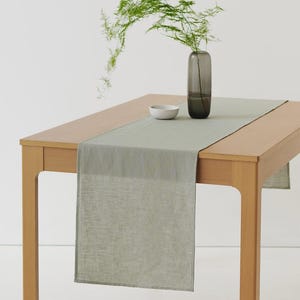 May include: A light sage green rectangular table runner made of a woven fabric draped over a light-toned wooden table. A small white ceramic bowl and a tall, slender, translucent dark gray glass vase containing a green fern sprig sit on top.