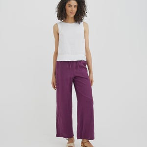 May include: A woman wearing a white sleeveless top and wide-leg purple pants. The pants have a drawstring waist. She is wearing beige sandals with a woven design. The background is a solid white.