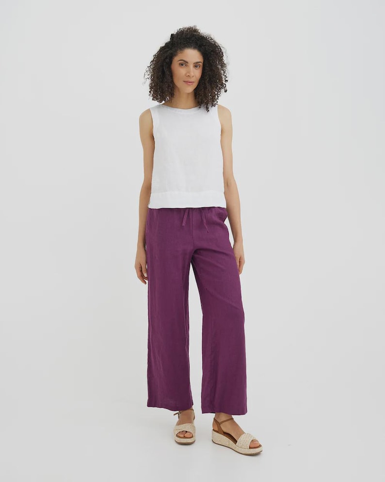 May include: A woman wearing a white sleeveless top and wide-leg purple pants. The pants have a drawstring waist. She is wearing beige sandals with a woven design. The background is a solid white.