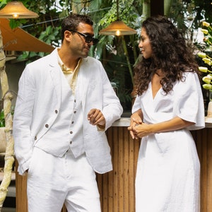 May include: A man and a woman wearing white linen clothing stand next to each other. The man is wearing a white linen suit with a vest and the woman is wearing a white linen dress. They are both looking at each other.