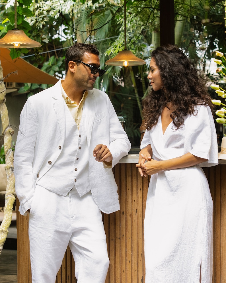 May include: A man and a woman wearing white linen clothing stand next to each other. The man is wearing a white linen suit with a vest and the woman is wearing a white linen dress. They are both looking at each other.