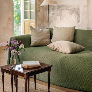 May include: A green sofa with a textured waffle-knit fabric cover, featuring three beige square throw pillows. In front of the sofa, two dark brown wooden nesting tables hold a clear glass vase with purple and white flowers and two stacked books.