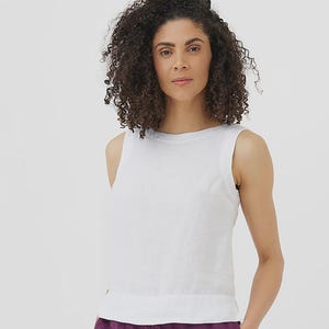 May include: A sleeveless white top with a round neckline. The top is made of a lightweight fabric. The model is wearing purple pants. The background is plain white.