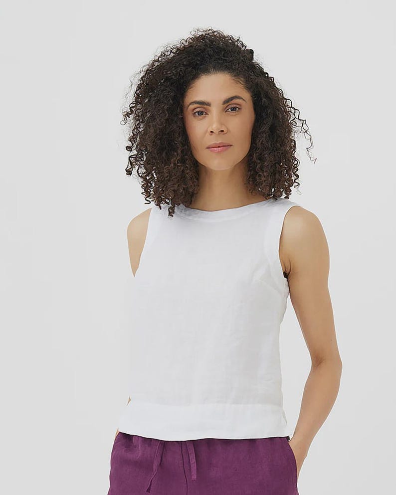 May include: A sleeveless white top with a round neckline. The top is made of a lightweight fabric. The model is wearing purple pants. The background is plain white.