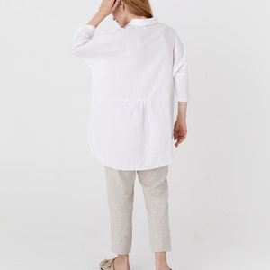 Pode incluir: Uma camisa de linho branca com um caimento relaxado e um comprimento ligeiramente mais longo. A camisa tem colarinho e mangas tr&ecirc;s quartos.