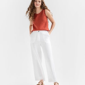 May include: A woman wearing a rust-colored sleeveless top and white linen pants. She is standing with her hands in her pockets and wearing brown sandals.