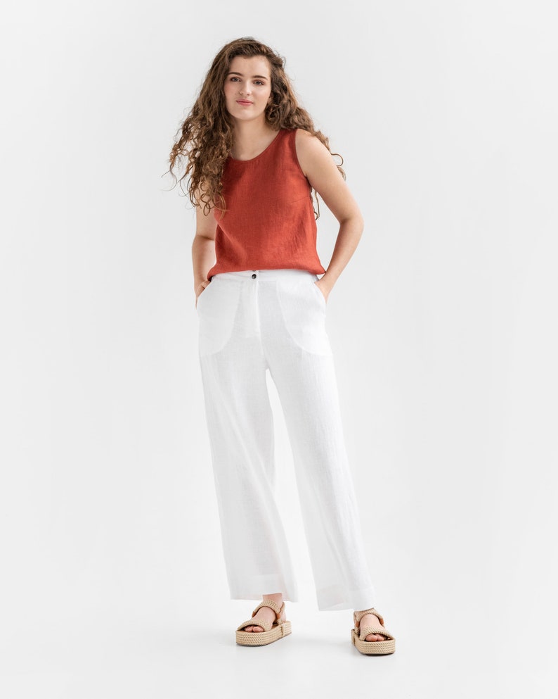 May include: A woman wearing a rust-colored sleeveless top and white linen pants. She is standing with her hands in her pockets and wearing brown sandals.