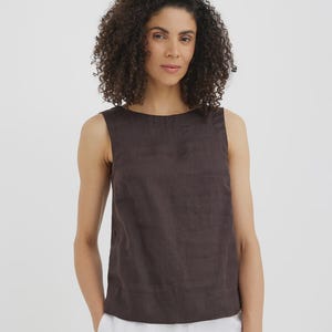 May include: A sleeveless, dark brown linen top with a round neckline. The top has a simple, minimalist design and is paired with white pants. The fabric appears to be slightly textured. The model is wearing the top.
