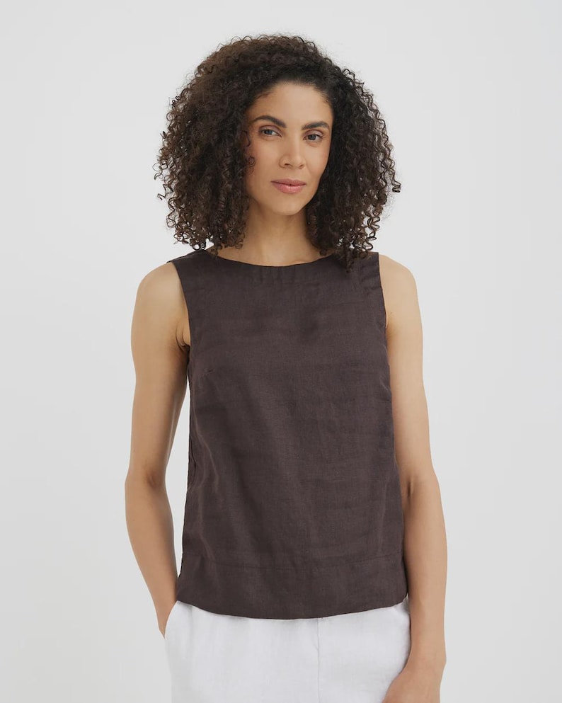 May include: A sleeveless, dark brown linen top with a round neckline. The top has a simple, minimalist design and is paired with white pants. The fabric appears to be slightly textured. The model is wearing the top.