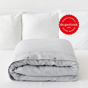 May include: A folded light gray duvet cover with a subtle pinstripe pattern and two round brown buttons at the top edge. A red circular badge in the upper right corner features the text: READER FAVORITE the good trade 2024 AWARD WINNER.