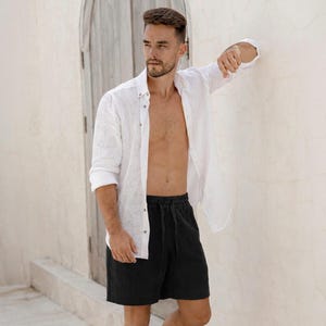 May include: A man wearing a white linen button-down shirt and black shorts with a drawstring waist. The shirt is unbuttoned, and the shorts are above the knee. The background is a light-colored wall and a wooden door.