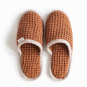 May include: A pair of brown waffle-knit slippers with a light beige trim. The slippers have a textured appearance and a soft, neutral color palette. A small label with text is visible on one slipper.