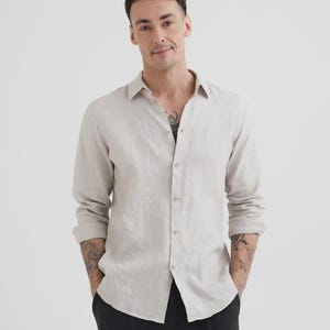 Pu&ograve; includere: Una camicia di lino beige chiaro con maniche lunghe e bottoni. La camicia ha un colletto classico e viene indossata con pantaloni neri. Le maniche sono arrotolate e la camicia &egrave; sbottonata in alto.