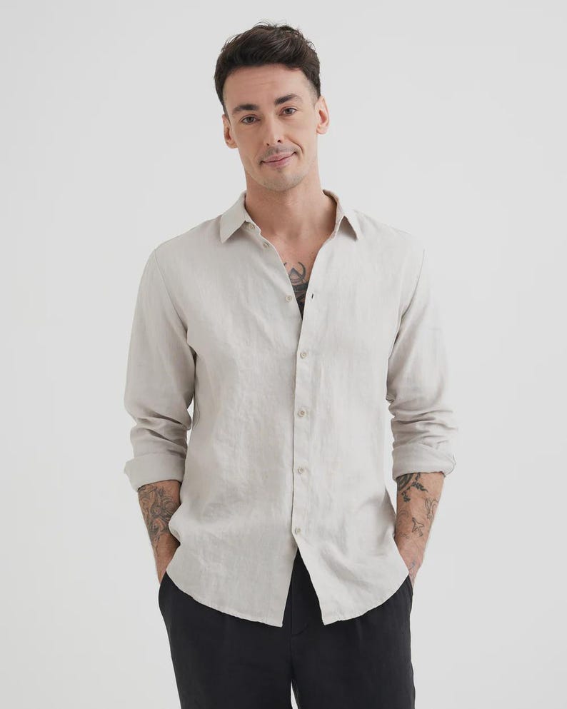 Pu&ograve; includere: Una camicia di lino beige chiaro con maniche lunghe e bottoni. La camicia ha un colletto classico e viene indossata con pantaloni neri. Le maniche sono arrotolate e la camicia &egrave; sbottonata in alto.