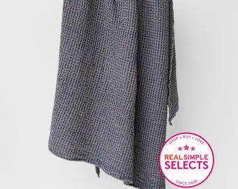 May include: A dark blue waffle-weave blanket hanging from a wooden rod. The fabric features a textured grid pattern. A circular pink and white badge in the bottom right corner displays the text: SHOP BUY LOVE, three stars, REAL SIMPLE SELECTS, SINCE 2000.