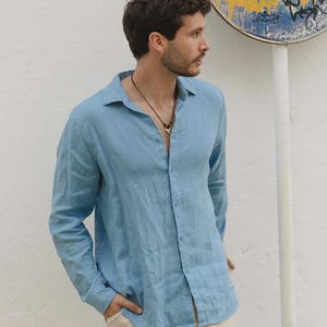 Pu&ograve; includere: Una camicia azzurra a maniche lunghe con un design button-down. La camicia &egrave; realizzata in un tessuto leggero e ha una vestibilit&agrave; rilassata. Il modello indossa una collana e pantaloni beige. Lo sfondo &egrave; un muro bianco con un cartello.