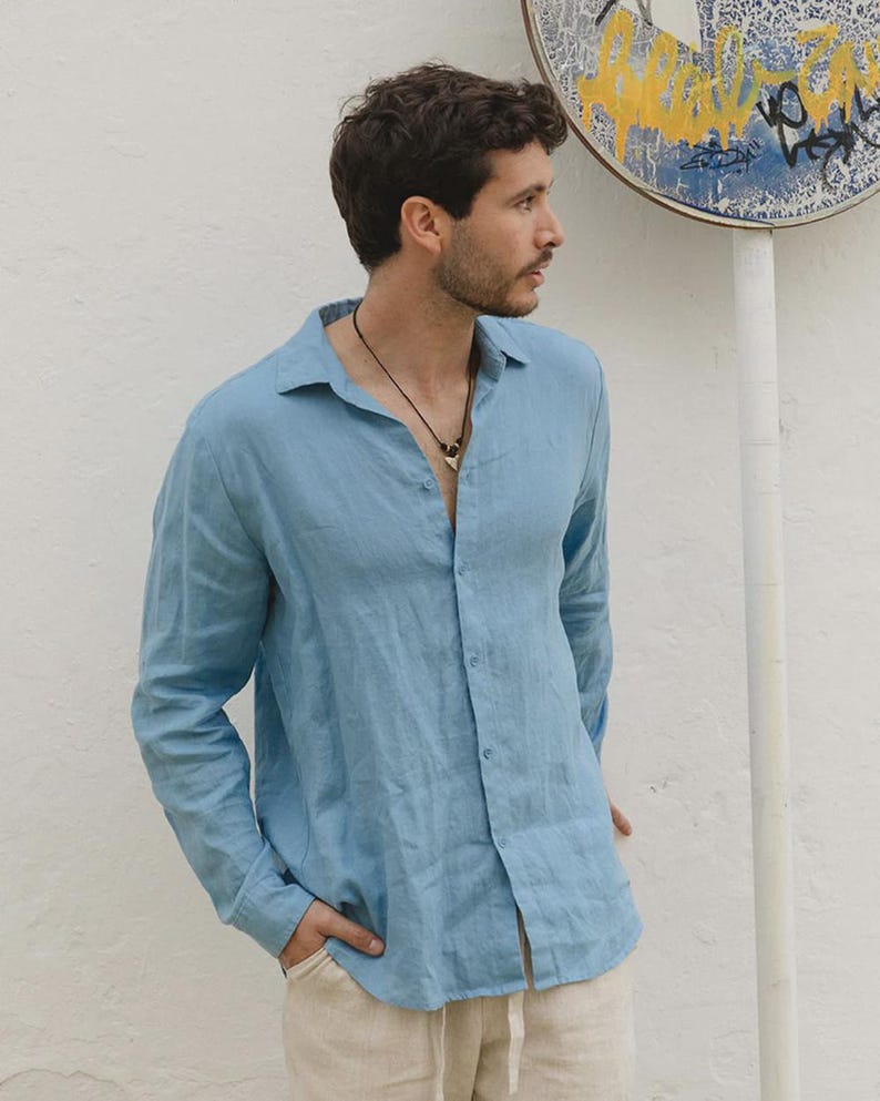Pu&ograve; includere: Una camicia azzurra a maniche lunghe con un design button-down. La camicia &egrave; realizzata in un tessuto leggero e ha una vestibilit&agrave; rilassata. Il modello indossa una collana e pantaloni beige. Lo sfondo &egrave; un muro bianco con un cartello.