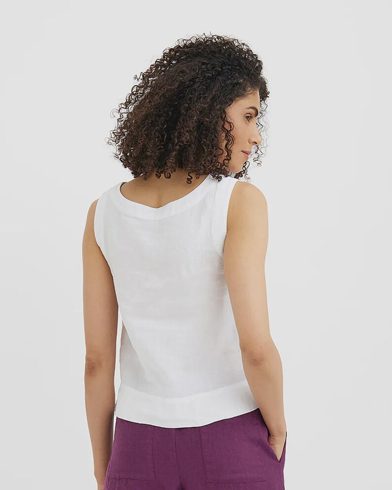 May include: A sleeveless white linen top with a scoop neckline. The top is paired with purple linen pants. The model is facing away from the camera, showcasing the back of the outfit. The fabric appears to be lightweight and breathable.