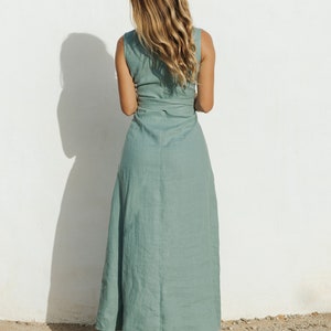 May include: A sleeveless sage green maxi dress featuring a wrap-style bodice with a tie waist detail. The dress has a long, flowing skirt that reaches the ankles and is constructed from a textured fabric, paired with tan platform sandals.