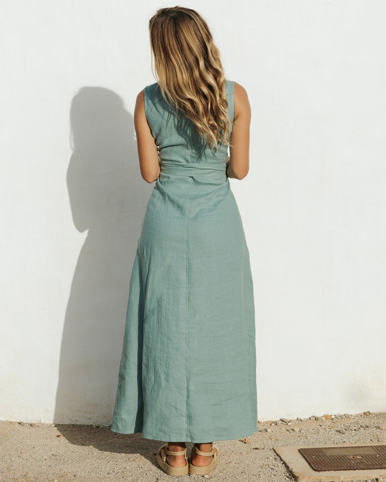 May include: A sleeveless sage green maxi dress featuring a wrap-style bodice with a tie waist detail. The dress has a long, flowing skirt that reaches the ankles and is constructed from a textured fabric, paired with tan platform sandals.