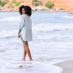 Pode incluir: Uma mulher vestindo um vestido de linho azul claro est&aacute; em uma praia de areia com ondas brancas e espumosas quebrando em seus p&eacute;s. O fundo mostra uma colina verde e &aacute;gua azul.