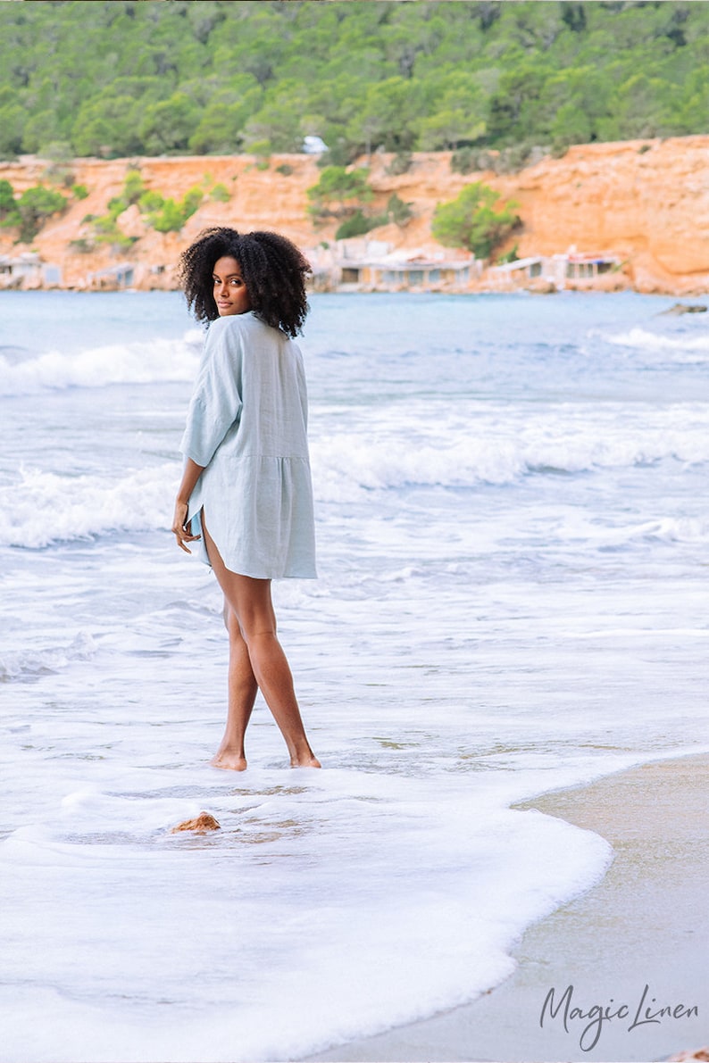 Pode incluir: Uma mulher vestindo um vestido de linho azul claro est&aacute; em uma praia de areia com ondas brancas e espumosas quebrando em seus p&eacute;s. O fundo mostra uma colina verde e &aacute;gua azul.