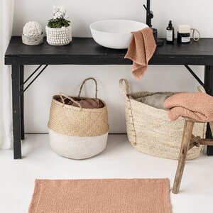 May include: A bathroom featuring a sink, two baskets, a stool, and a terracotta-coloured bath mat. The sink is white with a black tap. The baskets are made from natural materials and have white accents. The stool is made of wood and has a natural finish. The bath mat is made of a soft, absorbent material.