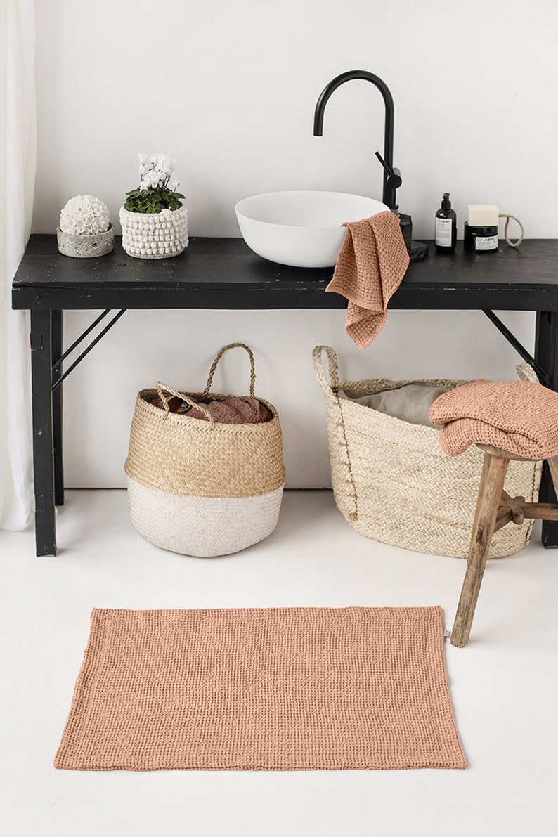May include: A bathroom featuring a sink, two baskets, a stool, and a terracotta-coloured bath mat. The sink is white with a black tap. The baskets are made from natural materials and have white accents. The stool is made of wood and has a natural finish. The bath mat is made of a soft, absorbent material.