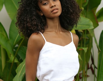 May include: A woman with curly dark brown hair is wearing a white tank top and a rust-coloured skirt with buttons. She is standing in front of a wall of green plants.